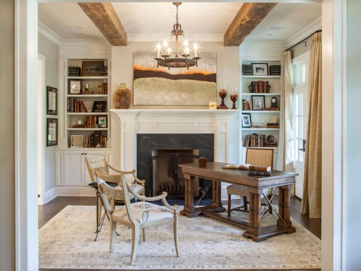 Traditional home office with a sturdy wood desk, fireplace, and rustic beams, illuminated by a chandelier