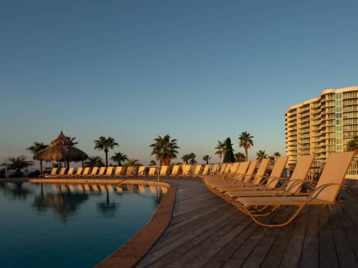 Resort poolside loungers lined up at sunset with palm trees and a condominium building in the background