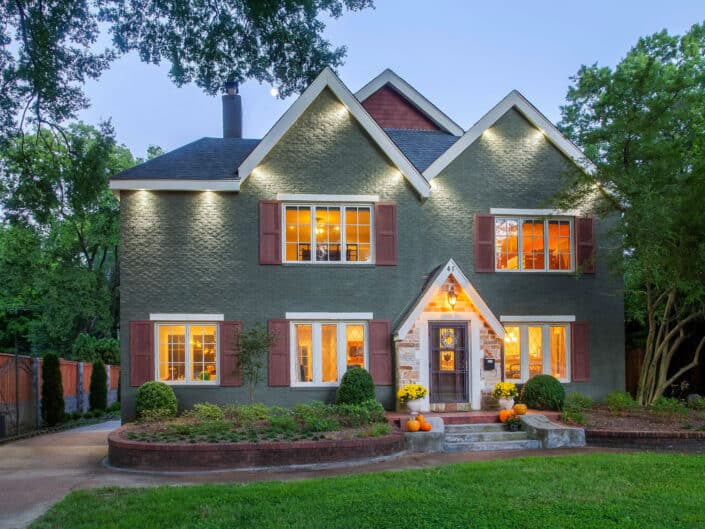 A charming two-story brick house adorned with festive autumn decorations, including pumpkins by the front door, surrounded by mature trees in the twilight.